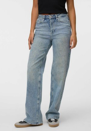 Persoon die lichtblauwe jeans met hoge taille en wijde pijpen draagt, met knoop en rits aan de voorkant, gecombineerd met zwart-witte sneakers.