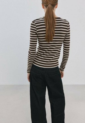 Ženska z dolgimi rjavimi lasmi, spetimi v čop, oblečena v dolgo rokavno majico z rjavimi in belimi črtami ter široke črne hlače, stoji pred enobarvnim ozadjem.