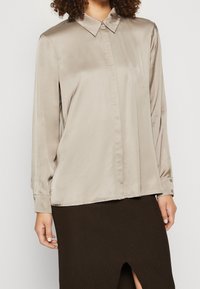 Beige Satin-Bluse mit langen Ärmeln, spitzem Kragen, Knopfmanschetten und geradem Schnitt. Glatte Textur mit subtiler Glanz.