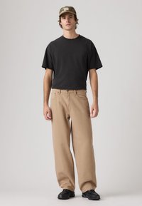 Μαύρο βαμβακερό T-shirt, μπεζ παντελόνι φαρδιάς γραμμής και μαύρα αθλητικά παπούτσια. Το μοντέλο φοράει καπέλο καμουφλάζ με έμπροσθεν τσιμπιδάκι λογότυπου.