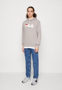 Sudadera gris con un logo de Fila en blanco y rojo, bolsillo frontal, combinada con jeans azules y zapatillas grises. Diseño sencillo y corte relajado.