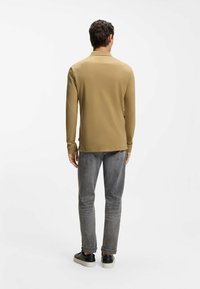 Koszulka polo z długim rękawem w kolorze khaki z kołnierzykiem, zestawiona z szarymi dżinsami i czarnymi trampkami. Minimalistyczny design, gładka faktura i dobrze dopasowany krój.