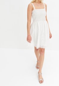 Vit sommarklänning med en figursydd smockad bodice, fyrkantig halsringning och justerbara axelband. Kjolen har detaljer med ögonlappar i spets längs fållen.