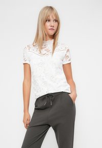 Bílý krajkový top s květinovými vzory, krátkými rukávy a vysokým výstřihem, doplněný tmavými kalhotami. Hladká textura, stylový design.