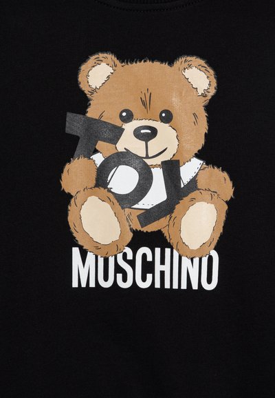 Juodas marškinėliai su pieštuku pavaizduotu meškiuku, laikantį žodį "ŽAISLAS" ryškiomis juodomis raidėmis, o žodis "MOSCHINO" atspausdintas žemiau baltomis raidėmis.