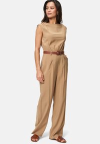Combinaison beige avec un col bateau, sans manches, taille cintrée et jambes larges. Fermée avec une ceinture marron et assortie à des sandales marron.