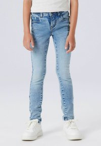 Přiléhavé světle modré džíny s hladkou texturou, pěti kapsami a jemným vyblednutím poblíž kolen, doplněné bílými teniskami.