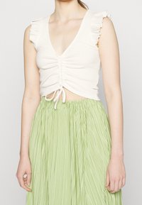 Bílý žebrovaný crop top s volánovými rukávy, s výstřihem do V a centrálním nařasením, zkombinovaný se světle zelenou skládací sukní.