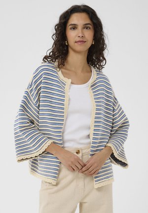 Žena s kučeravými tmavými vlasmi, oblečená v modro-bielom pruhovanom otvorenom kardigane cez biely top a béžové nohavice, stojaca pred jednoduchým pozadím.