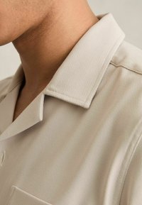 Camisa color beige claro con un patrón de espiga, que presenta un cuello puntiagudo y un pequeño bolsillo en el lado izquierdo. Textura suave y corte ajustado.