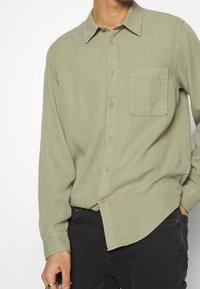 Camisa verde claro de manga larga con diseño de botones, cuello y un bolsillo en el pecho. Hecha de una tela suave con una textura lisa.