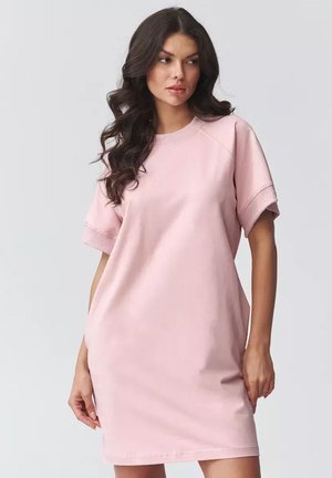 Kobieta z długimi, falującymi ciemnymi włosami, ubrana w luźną, pastelowo-różową sukienkę z krótkim rękawem, stojąca na tle jednolitego, jasnoszarego tła.