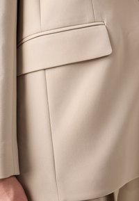 Beige blazer featuring smooth fabric, structured design, a hidden pocket, and clean lines. The texture appears textured but refined.