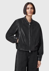 Giubbotto bomber in pelle nera con zip frontale, polsini a coste e colletto a banda. Presenta una superficie liscia e dettagli di cuciture curve.