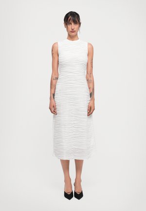 Witte midi-jurk met een textuur, gerimpeld ontwerp. Mouwloos, met een hoge halslijn. Gecombineerd met zwarte pumps met een spitse neus.