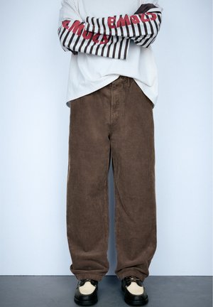 Image recadrée d’une personne debout, les bras croisés, portant une chemise blanche à rayures, un pantalon ample en velours côtelé marron et des chaussures noires.