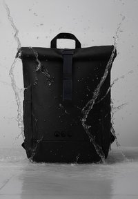 Schwarzer wasserdichter Rucksack mit Handgriff oben und Schnalle, Wasser spritzt umher vor einem schlichten grauen Hintergrund.