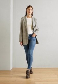 Blazer à carreaux en gris et beige, porté sur un haut blanc, associé à un jean skinny bleu et des chaussures à lacets marron, debout sur un sol en bois.