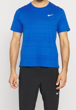 Homme portant un t-shirt Nike bleu à manches courtes et un pantalon Nike noir, debout devant un fond uni.