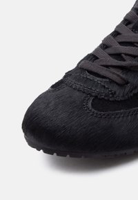 Μαύρο αθλητικό παπούτσι με υφή επάνω μέρος από γούνινο υλικό, ραμμένες επικαλύψεις και χοντρές μαύρες κορδόνια σε σκούρα λαστιχένια σόλα, εμφανιζόμενο από μπροστινή πλευρική γωνία.