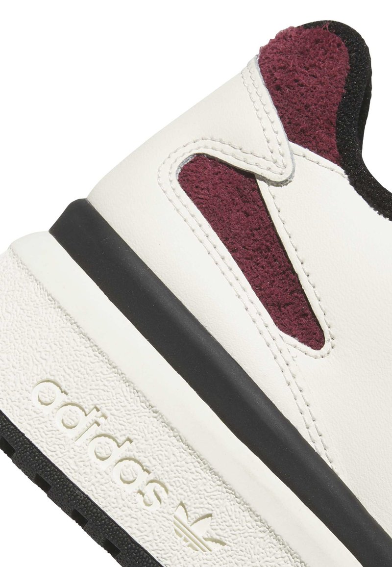 Baskets blanches en cuir texturé, avec des accents en daim bordeaux et une semelle noire. Elles présentent des détails cousus et un logo de marque sur le côté.