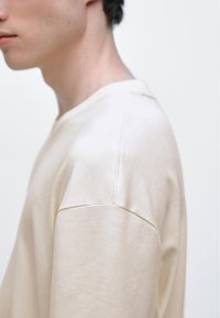 Beige t-shirt med rund halsringning, tillverkad av mjuk bomull med en avslappnad passform, som har något sänkta axlar och synliga sömmar.