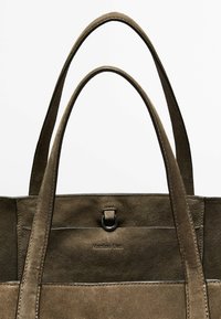 Bruine suède tote tas met twee lange handvatten, voorzien van een metalen ring en een geperst logo aan de voorkant. Zachte textuur en rechthoekige vorm.