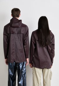 To personer bruker matchende mørk burgunder, vanntette jakker med hetter og klaffdesign, sammen med kontrastfargede, blanke og beige bukser.