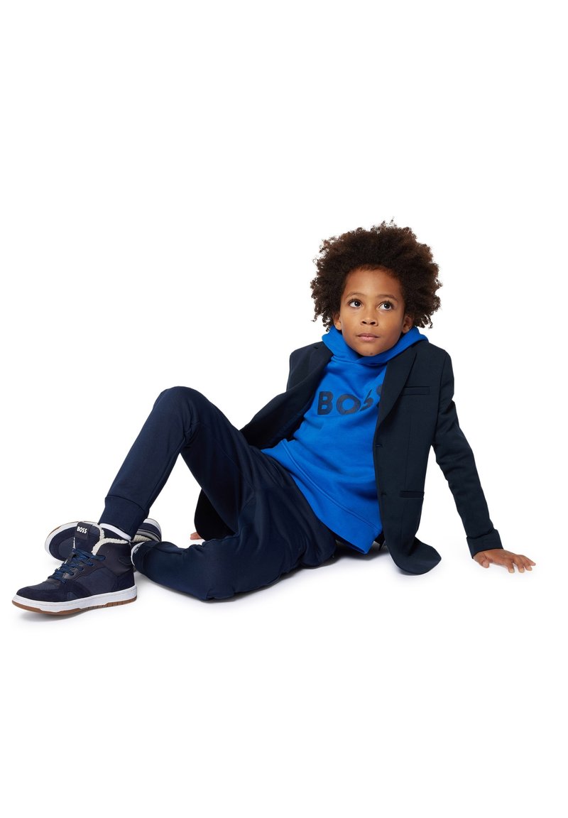 Veste de costume navy sur un hoodie bleu avec le logo "BOSS", associée à un pantalon de survêtement navy et des baskets. Modèle assis avec des cheveux bouclés.