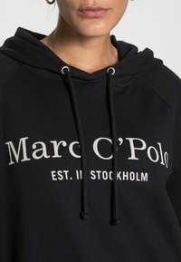 Sudadera negra con cordón, que presenta un gran texto en blanco: "Marc O’Polo EST. EN ESTOCOLMO." Tela suave y detalles cosidos en las mangas.