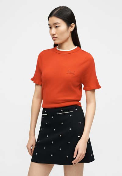 Γυναίκα με μακριά σκούρα μαλλιά, που φοράει κόκκινη μπλούζα με κοντά μανίκια και μικρό έμβλημα σκύλου, και μαύρη φούστα με λευκά σχέδια από χάντρες.