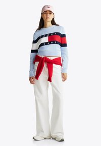 Jasnoniebieski sweter z dzianiny z czerwonymi, granatowymi i białymi poziomymi paskami, białe spodnie o szerokich nogawkach, czerwona bluza zawiązana w talii, różowa czapka.