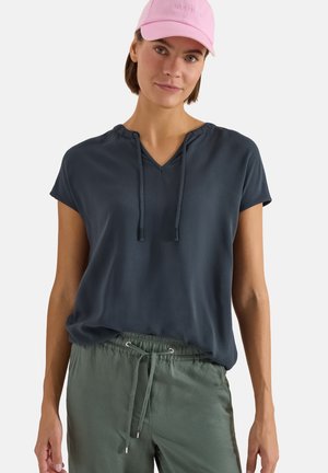 Femme portant une casquette rose, un haut bleu marine à manches courtes avec col en V et liens, et un pantalon vert à cordon, debout devant un fond blanc.
