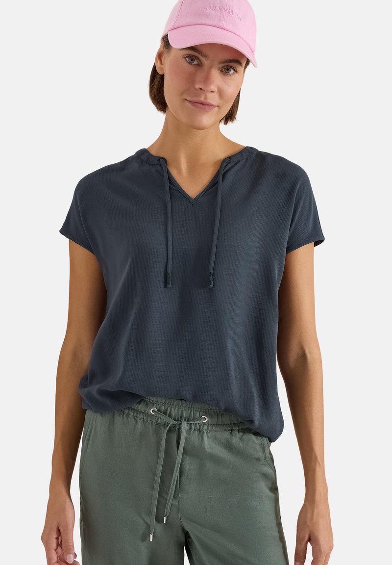 Femme portant une casquette rose, un haut bleu marine à manches courtes avec col en V et liens, et un pantalon vert à cordon, debout devant un fond blanc.