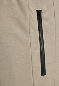 Beige tkaninski joggerji imajo stransko žep z črnim zadrgo, kar poudarja gladko teksturo in čiste šive.