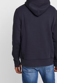 Sudadera azul marino con bolsillo tipo canguro, dobladillo y puños acanalados; presenta una textura suave y una capucha con cordón, combinada con jeans de color azul claro.