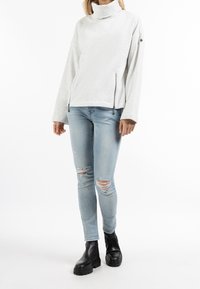 Suéter gris claro con un cuello alto, que cuenta con bolsillos laterales con cremallera y mangas sueltas. Combinado con jeans claros desgastados y botas negras de tobillo.