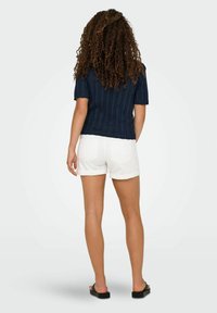 Marineblauw ribgebreid T-shirt met korte mouwen, gecombineerd met witte opgerolde shorts en zwarte sandalen, gezien vanuit de achterkant.