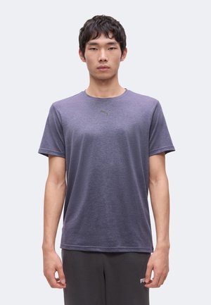 Junger Mann trägt ein schlichtes lila Puma-T-Shirt und dunkle Hose, steht mit neutralem Gesichtsausdruck und entspannter Haltung frontal.