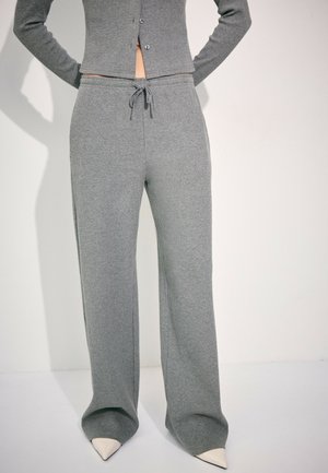 Person iført grå ribstrikkede vidde bukser med snøre i taljen og matchende knaplukket cropped top, stående med hænderne bag ryggen.