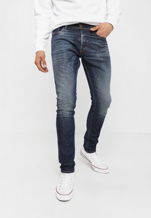 Mann trägt schlanke, dunkelblaue Jeans mit leichten Gebrauchsspuren, weiße Sneakers und ein weißes Langarmshirt, steht vor einem weißen Hintergrund.