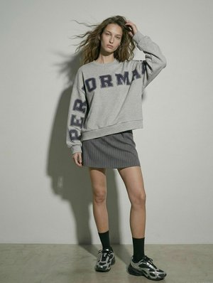 Mladá žena stojí, má na sebe oversized sivú mikinu s veľkým textom, krátku sukňu s jemnými pruhmi, čierne ponožky a robustné tenisky, ruka v vlasoch.