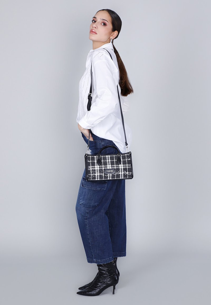 Donna con lunghi capelli castani, indossa una blusa bianca, jeans larghi blu e stivali col tacco neri, mentre porta una borsa a tracolla a scacchi bianco e nero.