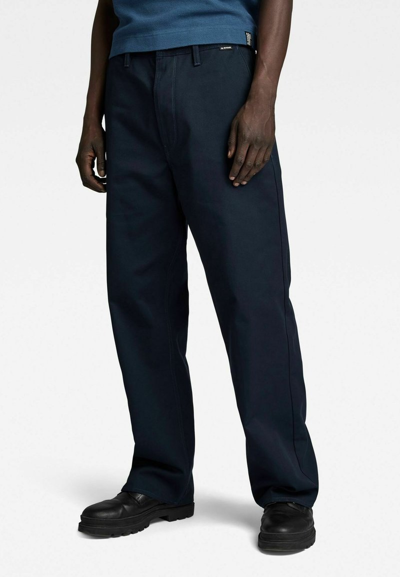 Donkerblauwe, losse broek van stevig materiaal, met een flat front en riemlussen. Draag met zwarte schoenen, detailsteekwerk is zichtbaar.
