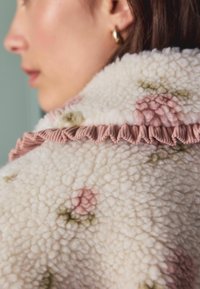 Fleecejakke med en tekstureret, fluffy overflade i creme, med pastelblomster i pink og grøn, samt en flæse i pink.