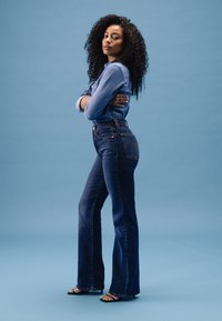 Jean flare en denim bleu avec taille haute et surpiqûres dorées contrastantes. Associé à un haut en denim à boutons, le tout sur un fond bleu.
