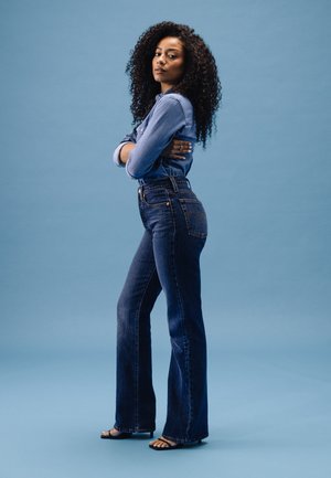 Blå denim flared jeans med høy livhøyde og kontrastfarget gullsting. Kombinert med en knapp-up denim topp, satt mot en blå bakgrunn.