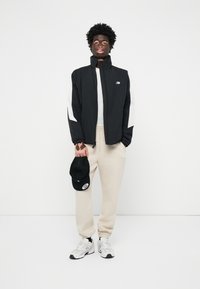 Svart jacka med vita detaljer, beige sweatpants och vita sneakers. Modellen håller en svart keps. Enkel, sportig design, avslappnad passform.