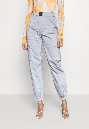 Žena v světle šedých kalhotách s elastickými manžetami, oranžovém vzorovaném topu s dlouhým rukávem a bílých sandálech na podpatku.