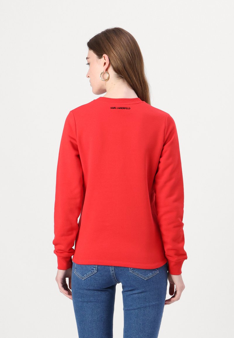 Roter Hoodie mit langen Ärmeln, Rundhalsausschnitt und Logo auf dem Rücken. Der Stoff wirkt glatt und wird mit engen blauen Jeans kombiniert.
